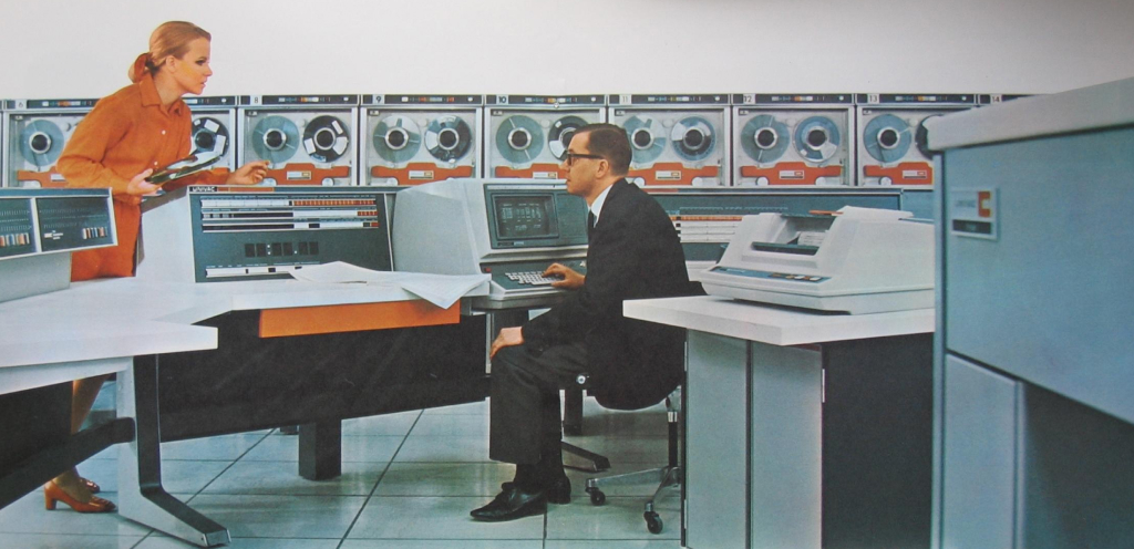 Image of a man in a suit sitting at a computer terminal from the 1960s.