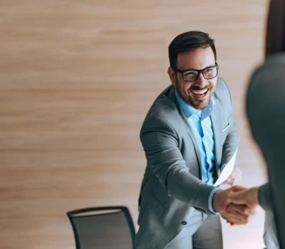 a smiling man in a suit reaching across a table to shake hands with another business professional