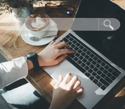 Photo of someone sitting at a desk with hands on a laptop keyboard with a transparent search bar overlayed at the top of the image.