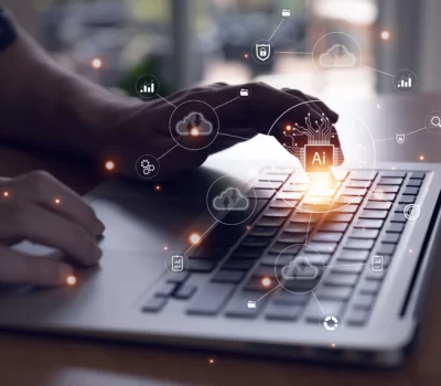 Person sitting at desk typing on a laptop, while small overlays of cloud and AI icons float on the screen, loosely connected to each other